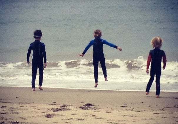 three children walk into the ocean wearing wetsuits