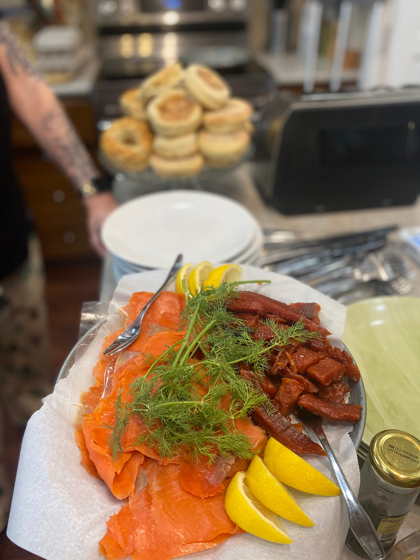 A platter of lox and maple-candied smoked salmon with sprigs of dill and wedges of lemon. In the background are a stack of plates and a pile of bagels and English muffins on a cake stand.