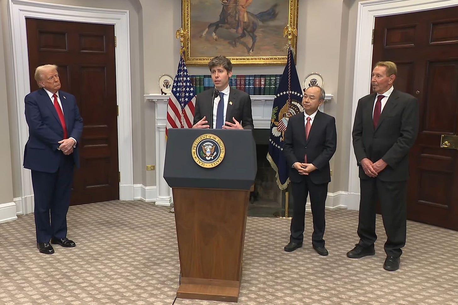 Donald Trump standing off to the side while OpenAI CEO Sam Altman speaks behind a lectern at the White House.