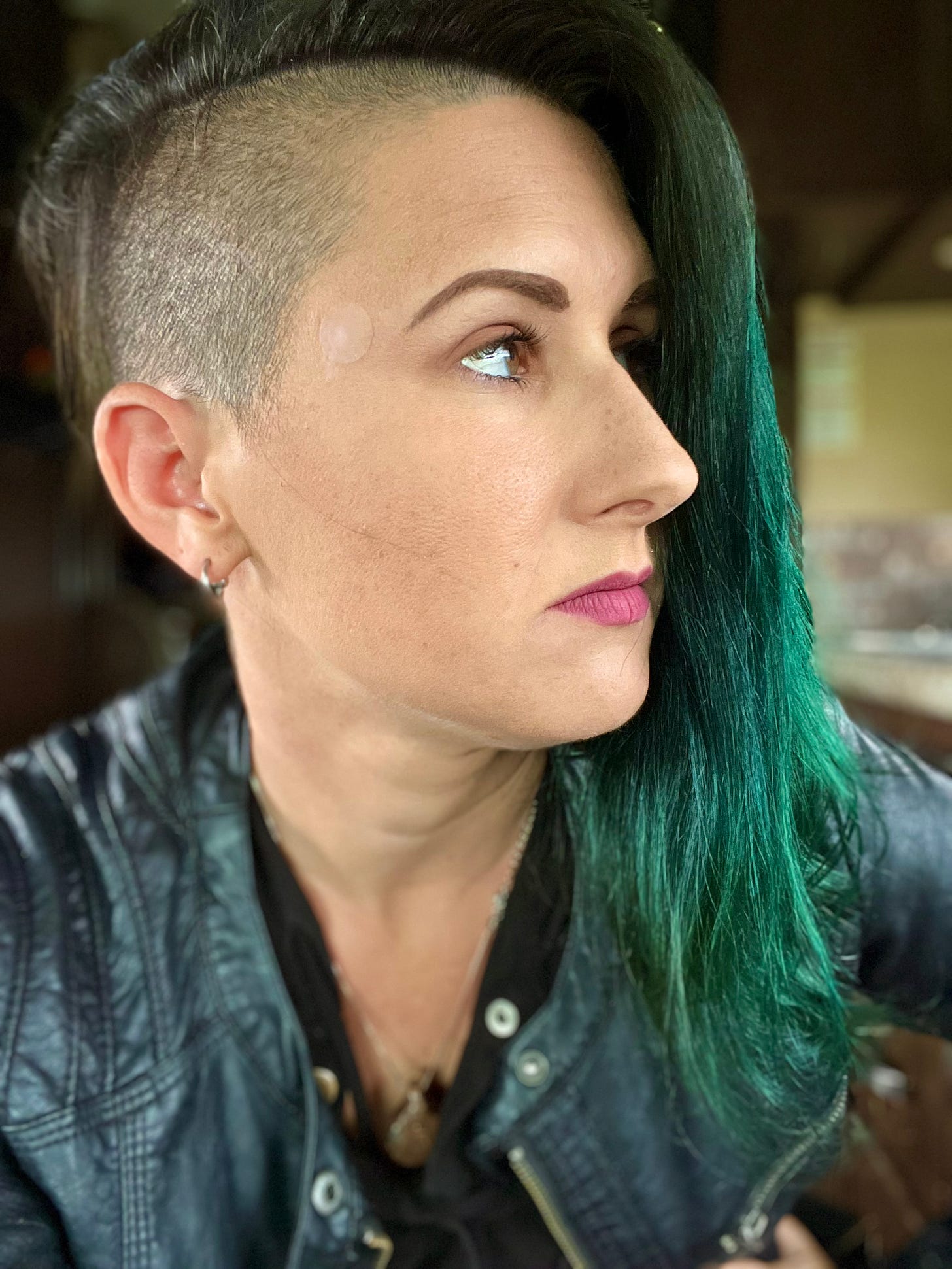 This is a photo of Lyric, with medium-length black hair with green ends and shaved sides, holding back tears. They are wearing a black leather jacket and can be seen inside their RV with the background blurred. 