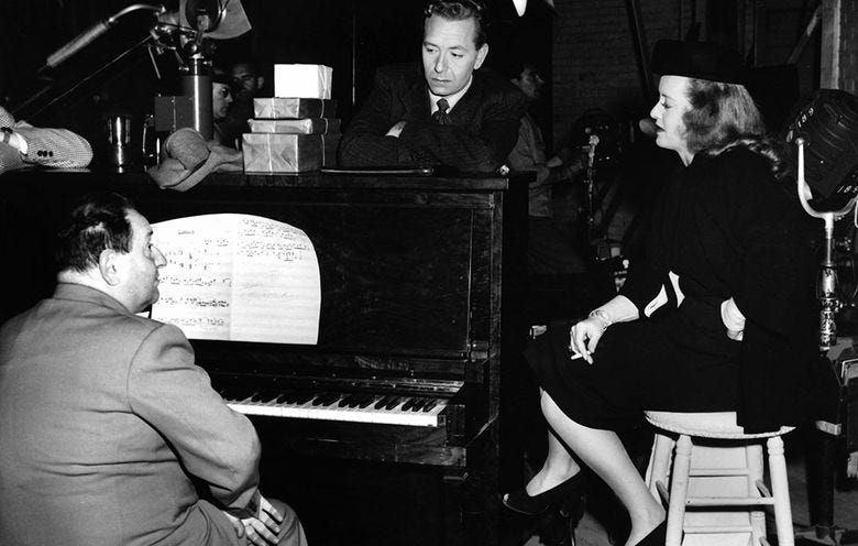 Black and white photo of a man sitting at a piano, another man standing next to the piano, and an woman seated in profile Black and white photo of a man sitting at a piano, another man standing next to the piano, and an woman seated in profile