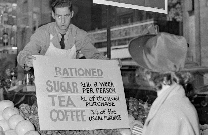 A 1940s store clerk holds a sign listing ration limits for sugar, tea and coffee