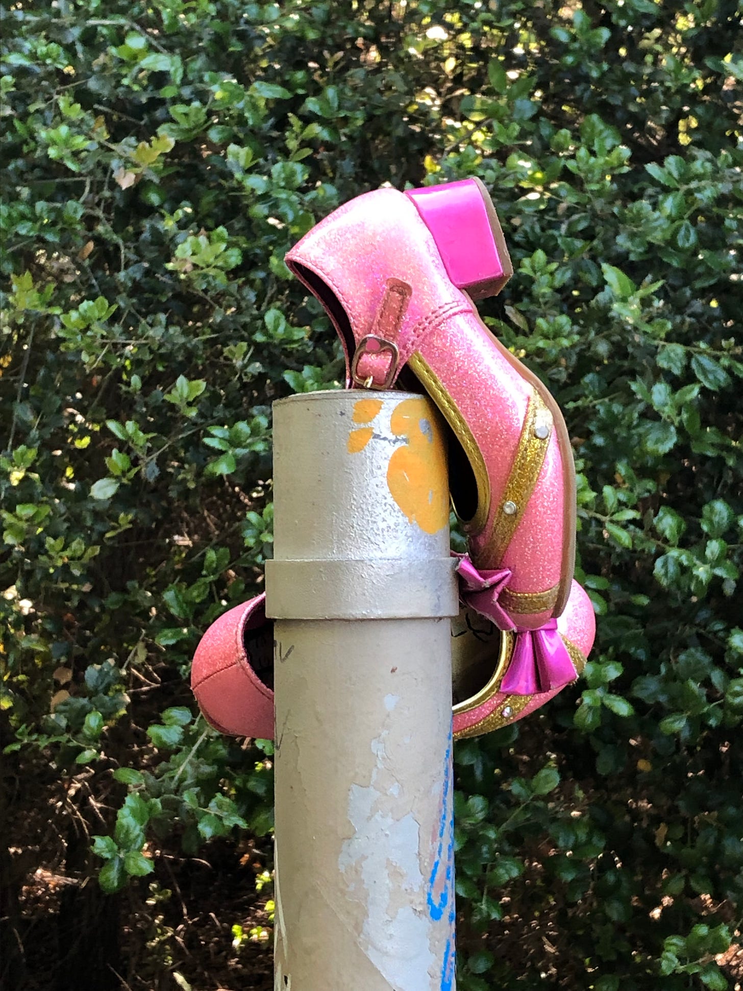 A pair of girls’ shoes hanging off the top of a metal post. The shoes are shiny, glittery pink with gold accents and a few rhinestones.