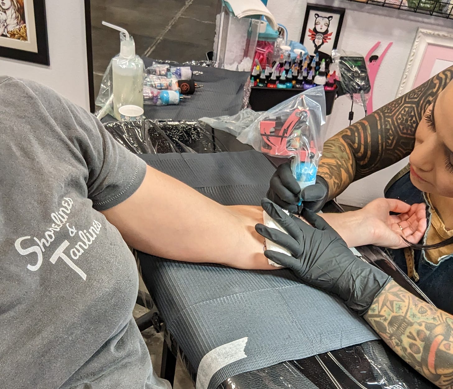 Joanna's arm being tattooed by Alex Strangler, a woman with tattooed arms and long, dark eyelashes