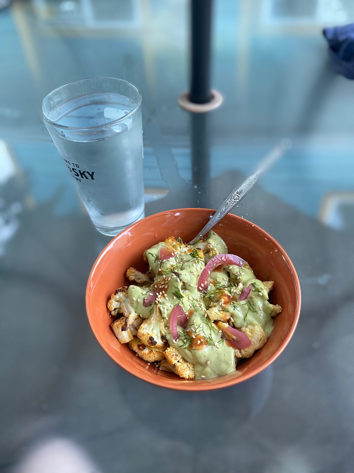 An orange bowl of quinoa, roasted, cauliflower, and tofu covered with green goddess dressing. On top are pickled red onion slices, sesame seeds, and dots of hot sauce. Next to the bowl is a pint glass of water with condensation on the outside.