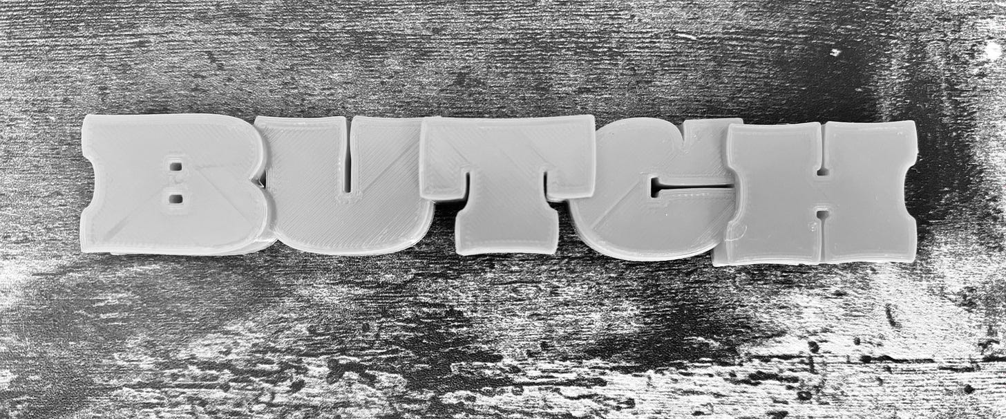 a 3d printed block text that says BUTCH against woodgrain in black and white