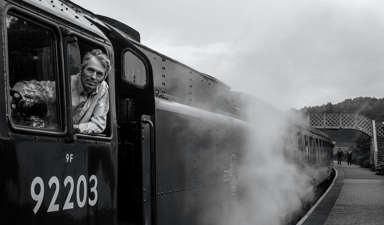 Schwarz-Weiß-Foto einer Dampflok von schräg vorne an einem Bahnhof. Ein Lokführer schaut aus dem Fenster nach vorne. (Foto: Lee Hull auf Unsplash)