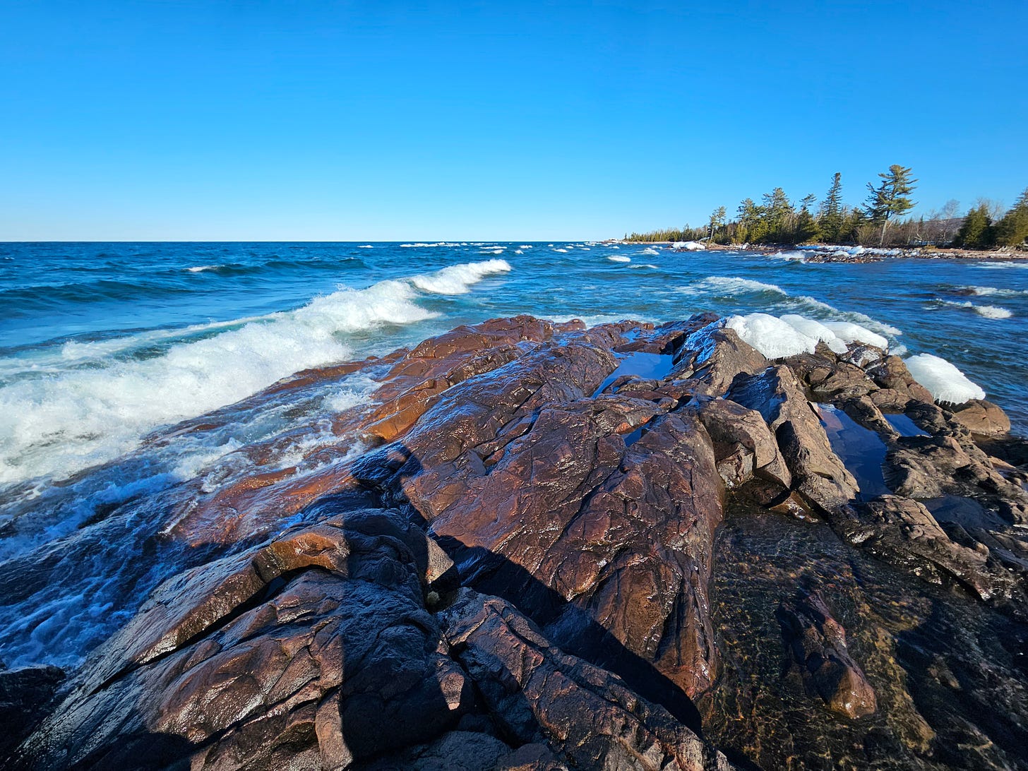 Emily's shadow stretches out across the ice- and snow-covered rocks of a stony outcrop into a turbulent and very blue Lake Superior.