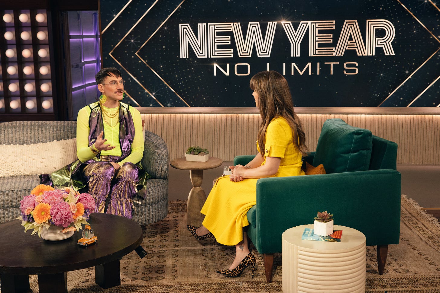 A photo from the set of The Kelly Clarkson Show. It’s a living room with cozy seating, bright flowers and a taxi cab snowglobe on a coffee table. Kelly Clarkson is a white femme wearing a yellow dress and leopard heels talking to Kevin Gotkin, a white person with a mustache wearing neon green and shimmering purple who looks like a Professor of Nightlife Studies at Shiz University from Wicked. On a screen in the background, it says “New year, no limits.”