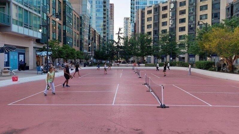 Picture from Change.org petition of a red pickleball court with about six set-up nets all being utilized. This is in the center of a cluster of apartment buildings, with the courts being what appears to be 25 to 30 feet away from some of the buildings. In the background small trees looms along the sidewalk and people are on benches on the sidewalk just beyond the courts. Picture from Change.org petition of a red pickleball court with about six set-up nets all being utilized. This is in the center of a cluster of apartment buildings, with the courts being what appears to be 25 to 30 feet away from some of the buildings. In the background small trees looms along the sidewalk and people are on benches on the sidewalk just beyond the courts.