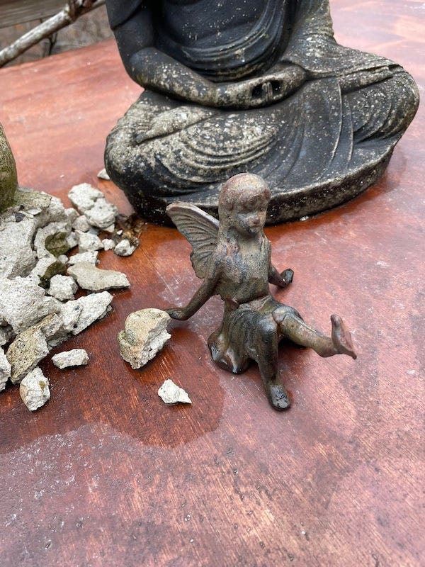Photo of disintegrating gargoyle and intact Buddha. Betty the wood nymp, who looks like a girl of perhaps ten years old, wearing a summer dress, has joined them. She is much smaller than the other two, & made of cast metal. She is seated, leaning back on her hands, with her left leg kicking up in the air. 