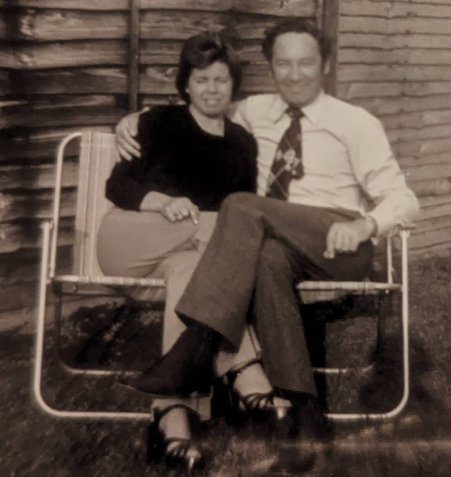 A man and woman sit on deckchairs in a small garden, arms and legs intertwined