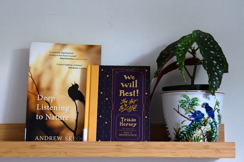 Library copies of ‘Deep Listening to Nature’ by Andrew Skeoch and ‘We Will Rest! The Art of Escape’ by Tricia Hersey sit on a shelf, next to a begonia plant.