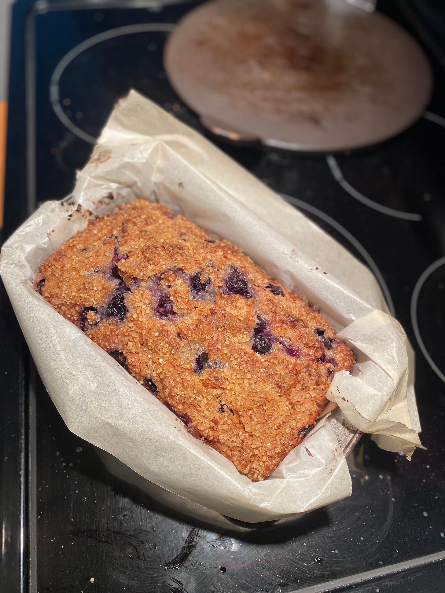 The blueberry loaf described above, in a loaf pan lined with parchment, resting on top of the stove. The top is coated in turbinado sugar and burst blueberries can be seen throughout.