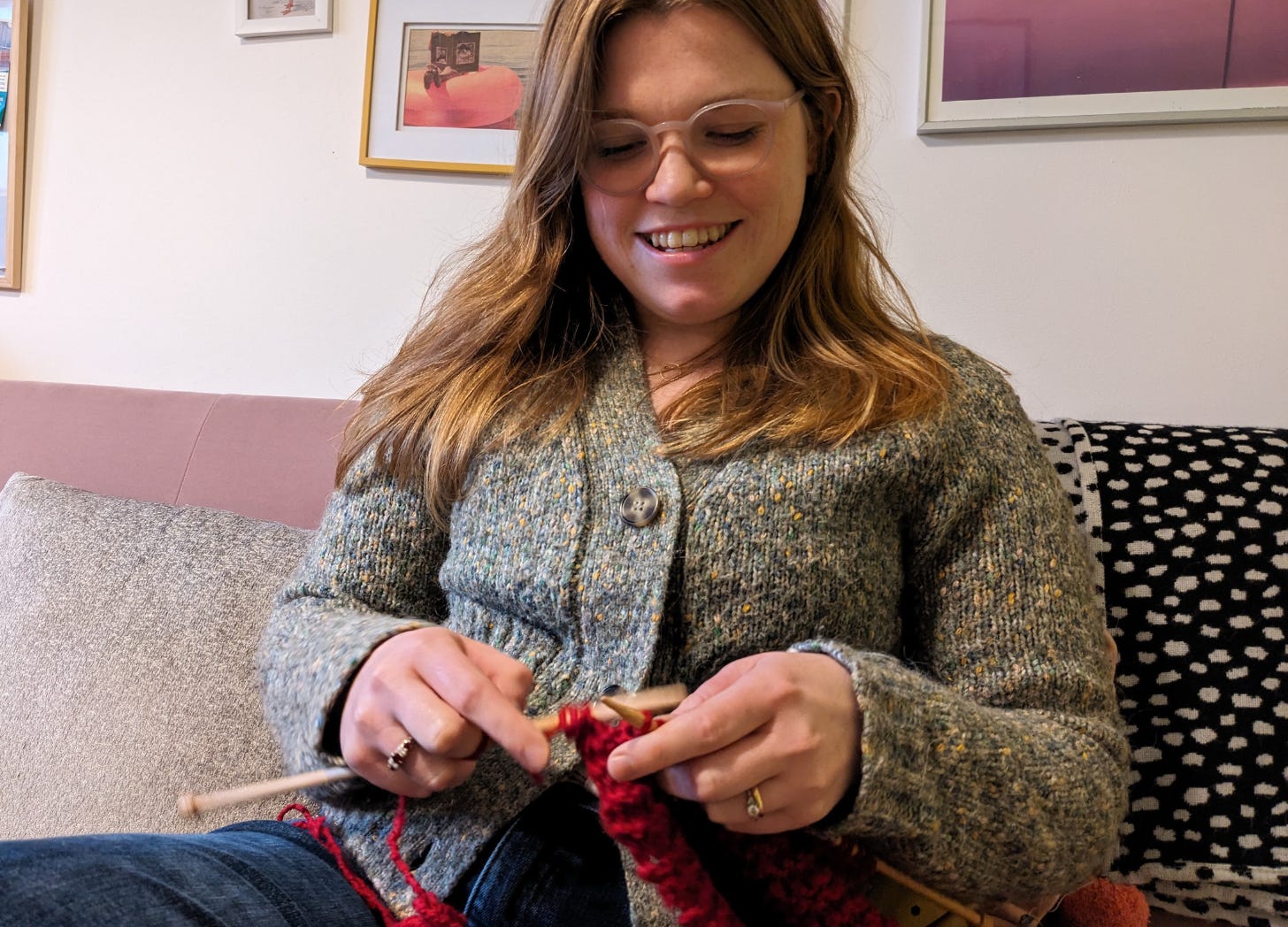 Libby Page sits on a sofa, wearing a grey jumper, knitting with red wool. She is smiling and looking down at her knitting. 