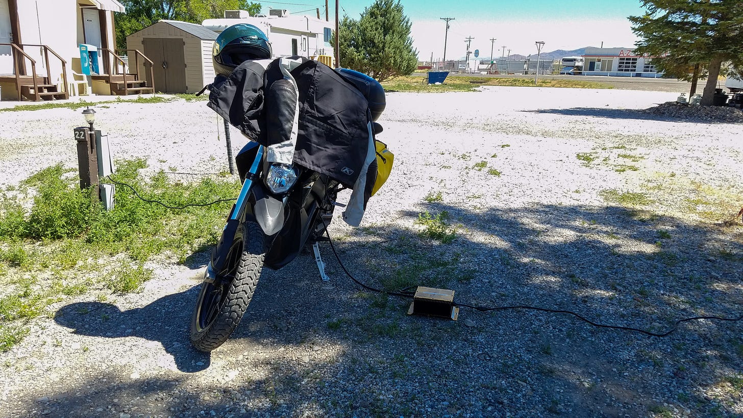 Photo of Zero electric motorcycle parked partially in the shade of a small RV park. There are patches of grass and weeds near the wooden pole on the left that contains one of the plugs that the electric motorcycle is plugged into. On the right, a cord frons from the motorcycle to a shoe-box sized transformer sitting in the shade on the ground. Another cord runs from the box to another plug to the right, just beyond the frame.