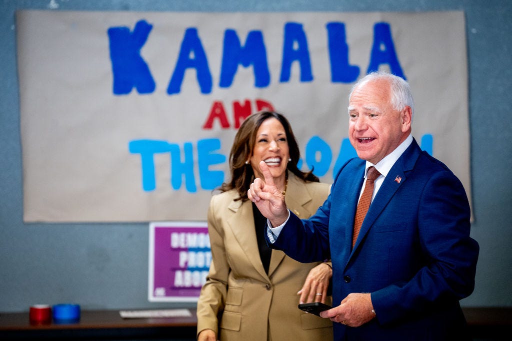 Vice President Kamala Harris and Minnesota Governor Tim Walz at a campaign appearance, in front of a hand-painted sign reading "Kamala and The Coach"