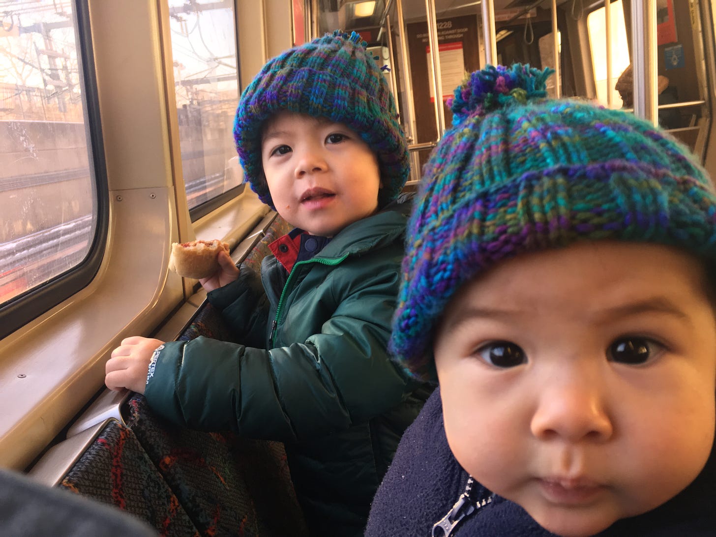 Baby Cass and toddler Blaise smile for a picture wearing knitted winter hats on an Orange Line MBTA train