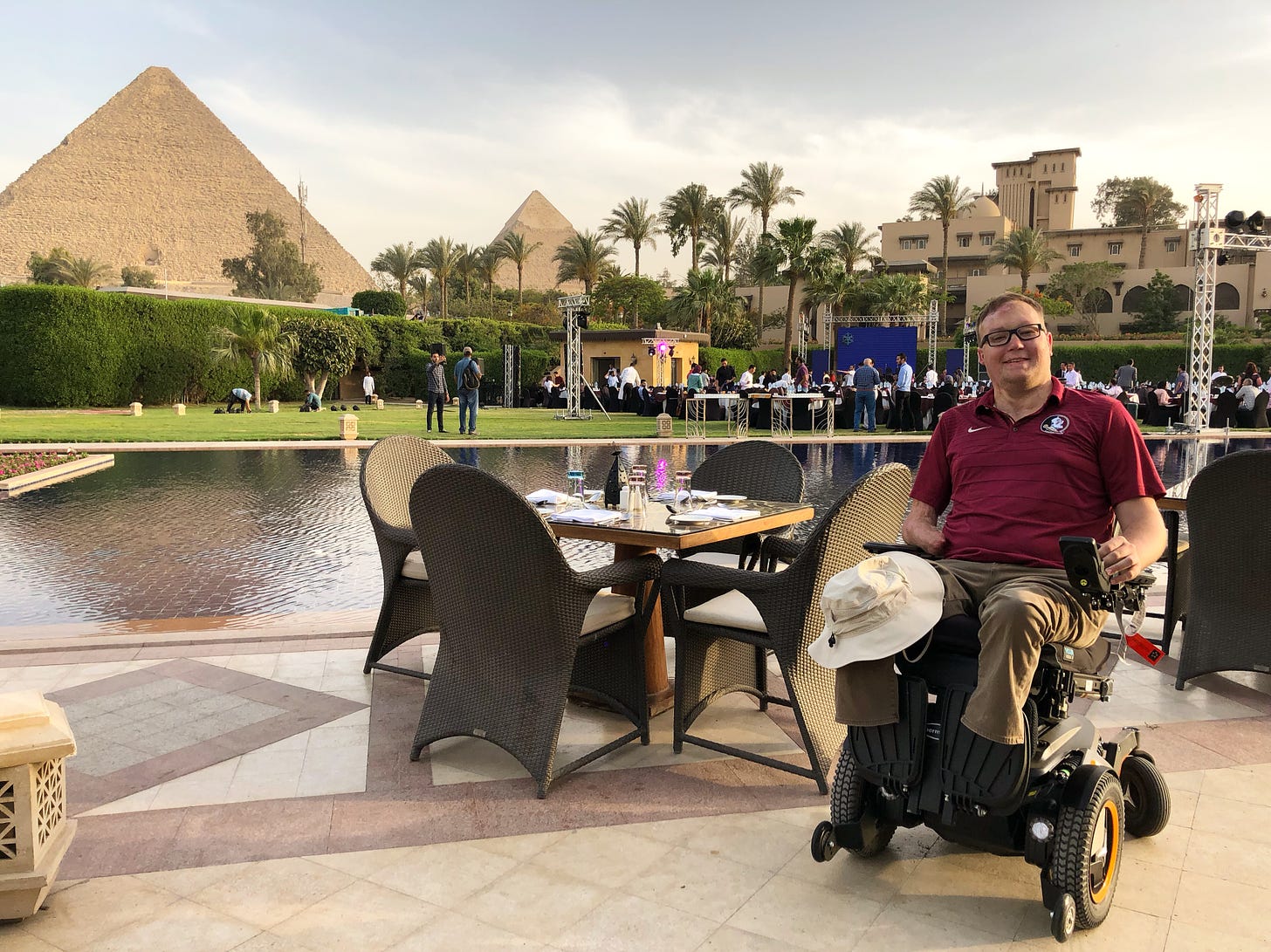 John seated in his power wheelchair at an outdoor table in front of an Egyptian pyramid.