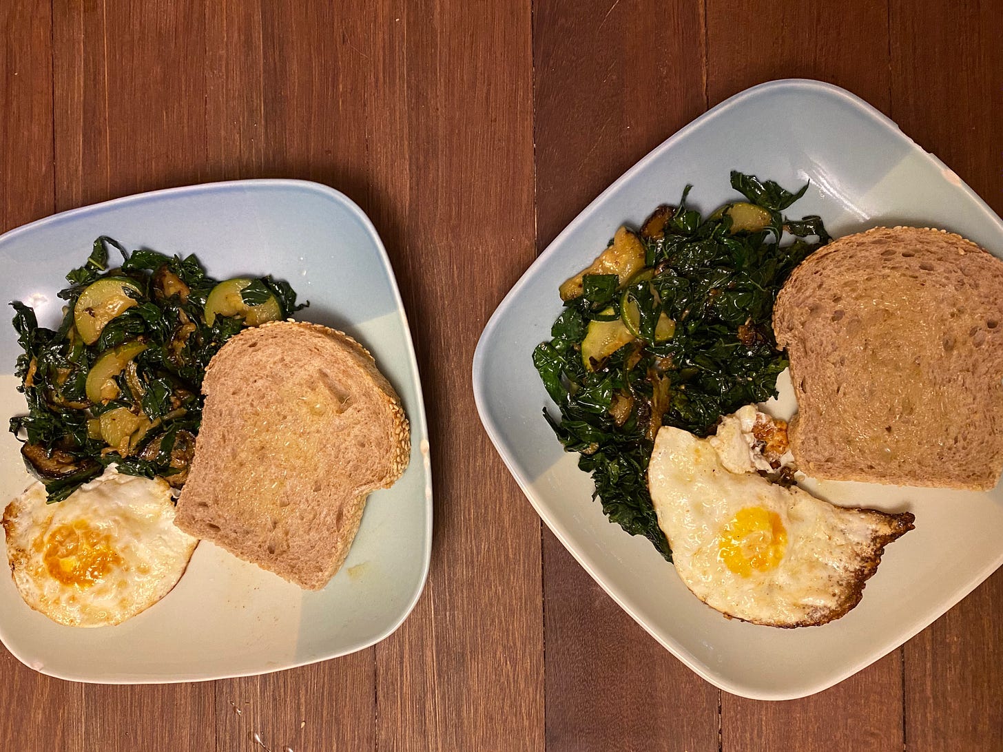 Two square plates on a wooden counter, each filled with a pile of kale and sautéed summer squash, a piece of butter toast, and a fried egg.