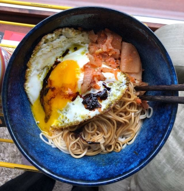 A dark blue ceramic bowl of noodles with a fried egg and kimchi on top, and spring onion soy sauce oil, and wooden chopsticks.