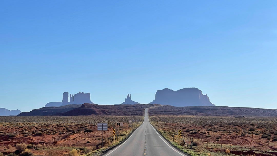 A highway disappears into the distance, where you can see the silhouettes of Monument Valley on the horizon.