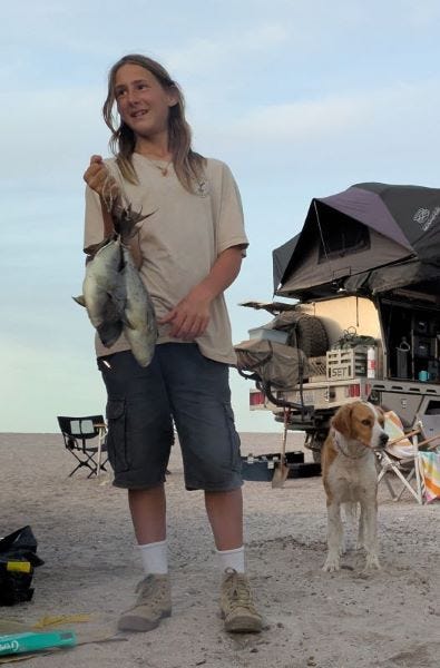 a teenage boy with long hair holds a string of fish he just caught while a dog watches nearby. in the background is a truck with a rooftop camper parked on the sand and assorted chairs and towels facing the long rays of sunset