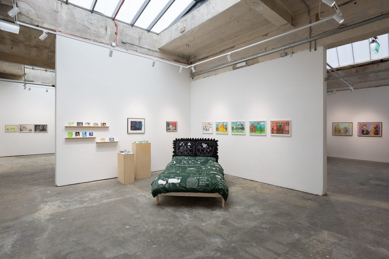 In a gallery with concrete floors, white walls, and natural light, works hang on the walls, on thin shelves, and a bed is in the middle of the gallery.