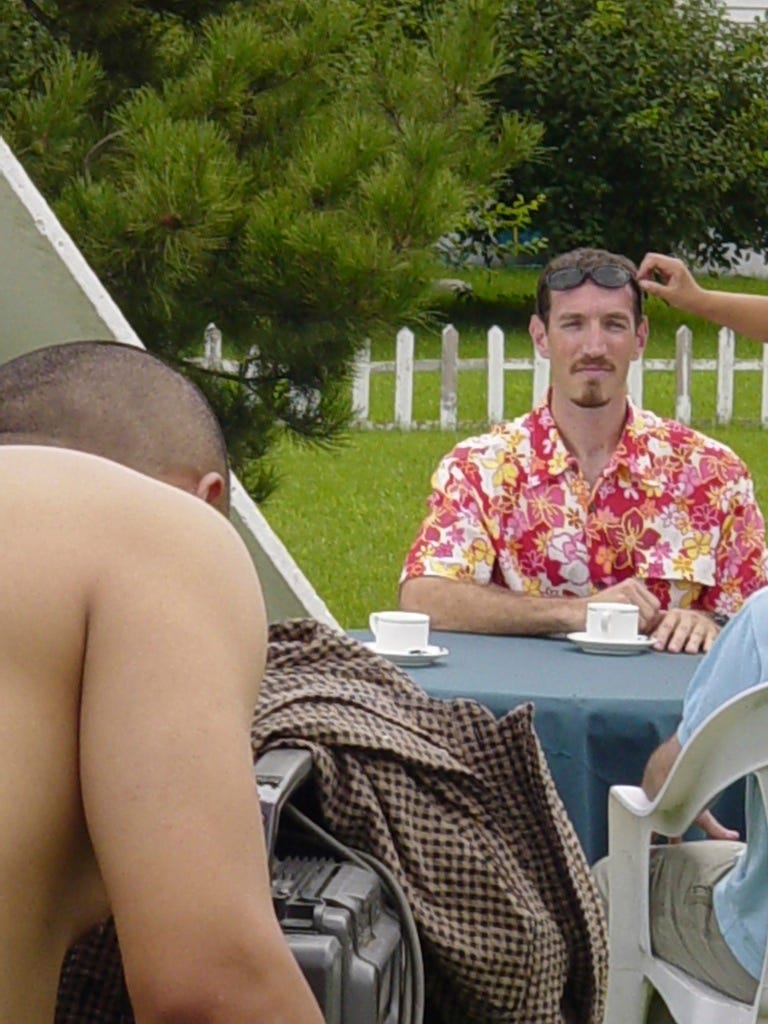 A cameraman lines up to film a goateed American man seated at a table on a lawn. The American is wearing a very loud Hawaiian print shirt and has sunglasses on his forehead. A hand reaches in from out of frame to adjust the glasses.