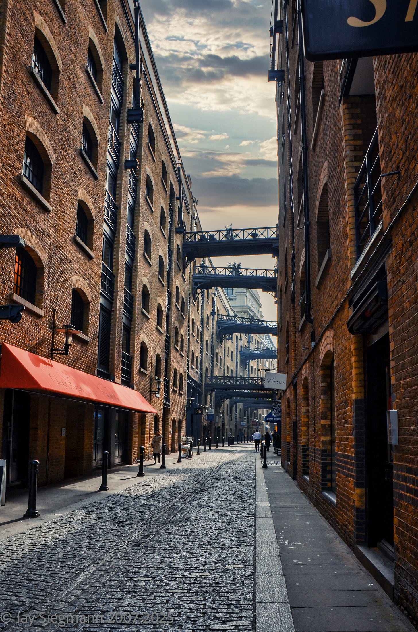 Historic industrial architecture of Butler's Wharf on Southbank, London, captured by Jay Siegmann. Cobbled streets stretch between brick warehouses, connected by cast-iron bridges, under a moody, cloud-streaked sky