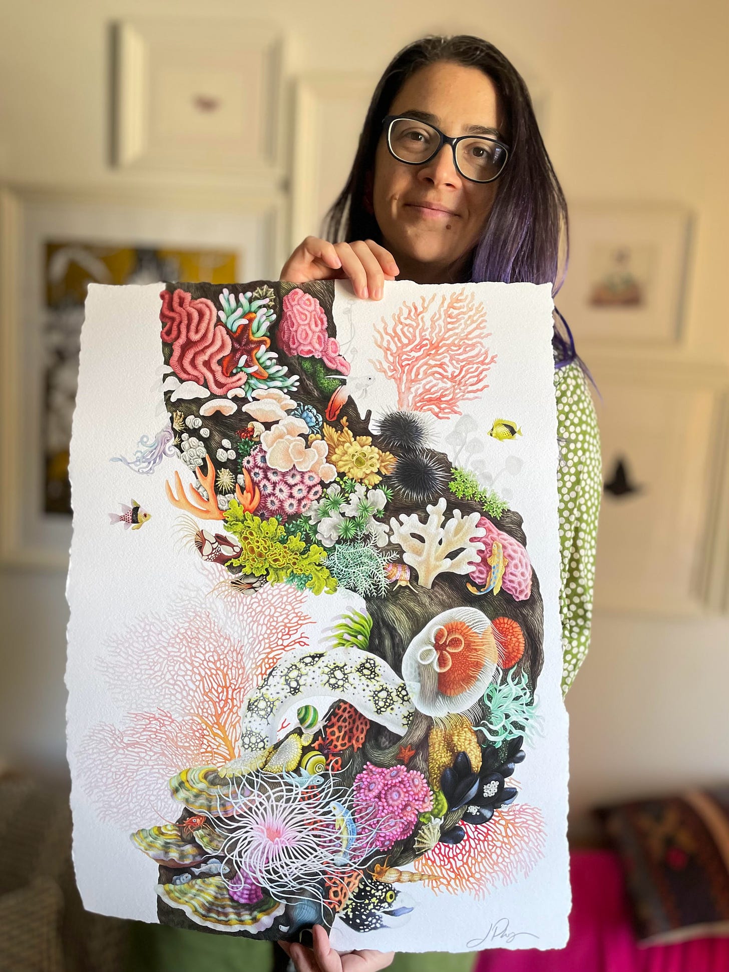 Lidija, a tan-skinned woman with brown eyes, purple hair, and glasses, holds one of her creations, a color illustration of a coral reef with fish, sea anemones, urchins, jellyfish, an octupus, and other sea creatures