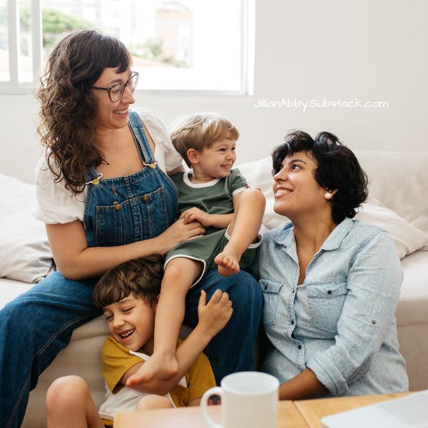 Lesbian couple in denim outfits sit on a white couch with their two children.