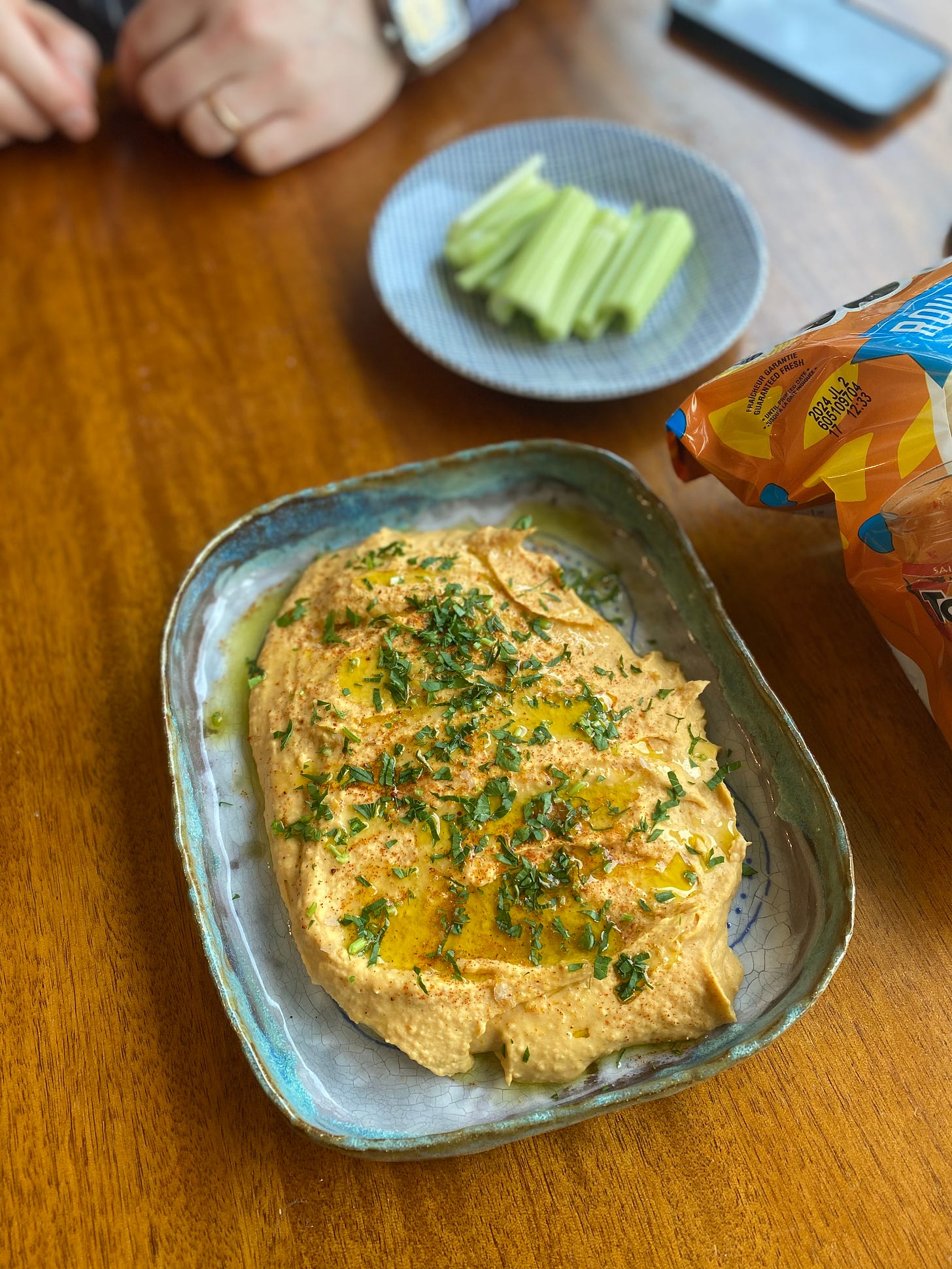 A light orange hummus dusted with spices and parsley, and drizzled with olive oil, in a blue ceramic dish. On a small plate are sticks of celery, and a bag of tortilla chips is just visible on the right side of the frame. Jeff's hands rest on the table in the upper left.