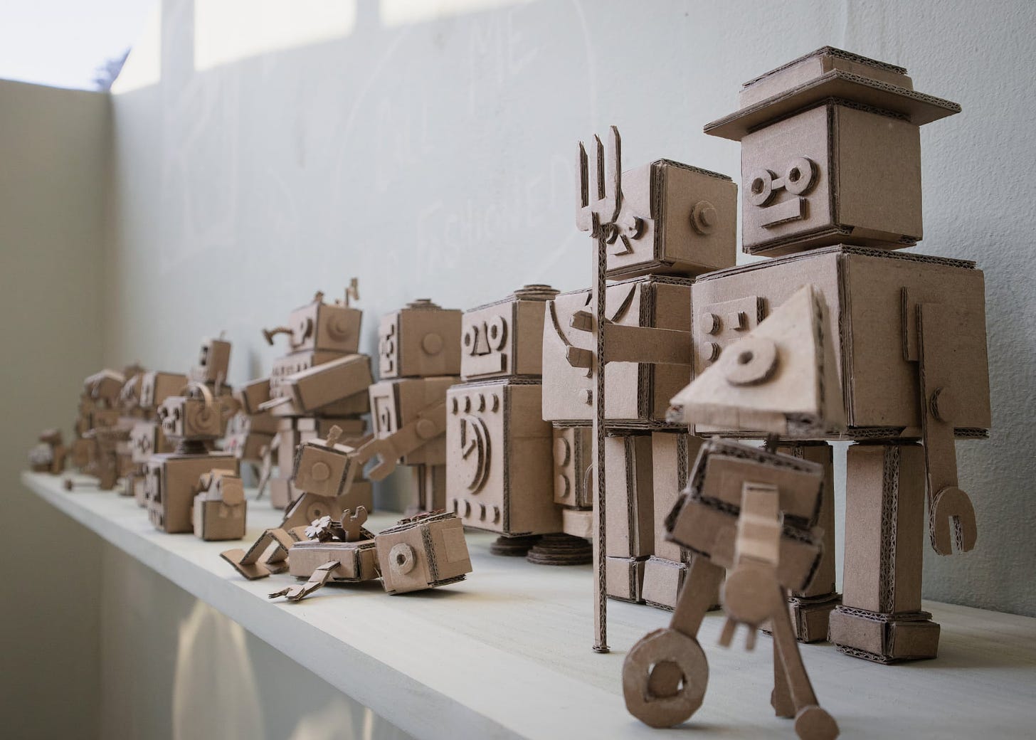  collection of cardboard robots by Beth Edgar displayed at Gray’s Degree Show. The robots are creatively crafted from corrugated cardboard, each with unique shapes and designs. They are arranged on a white shelf against a light wall, with shadows casting gently behind them. The robots vary in size and form, some resembling humanoid figures while others take on more abstract or mechanical shapes. The art installation showcases a playful and inventive use of recycled materials to create an assembly of robot figures.