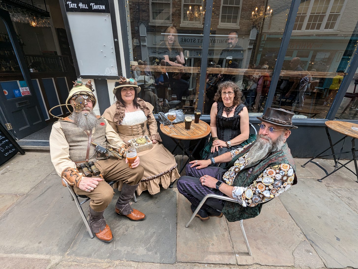 Four steampunkers sit around a table outside a pub, looking friendly