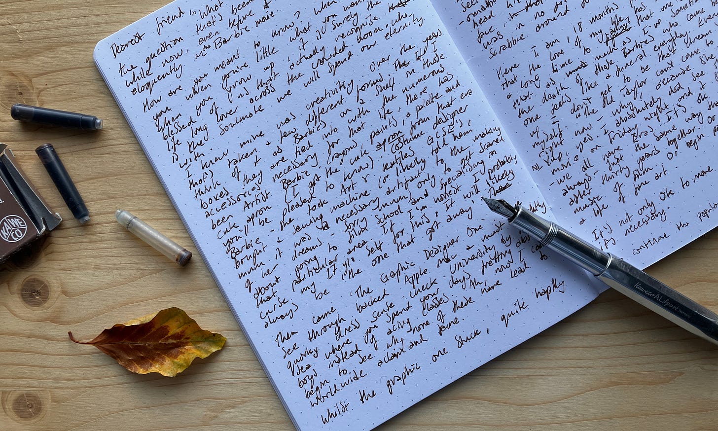 Image of notebook with draft letter on a desk with an empty pen cartridge some inky cartridges and an autumn leaf