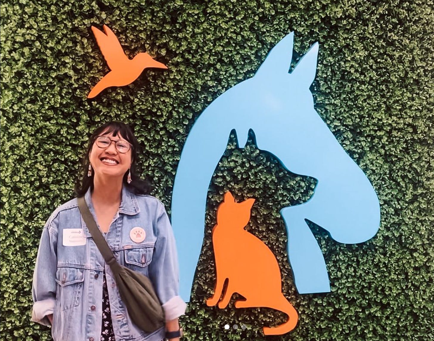 A smiling woman in glasses with a thick strapped purse slung over her jean jacket smiles broadly for the camera in front of a green ivy wall and wall attached art of an orange cat silhouette inside of a blue dog silhouette. An orange bird silhouette flies above against the green ivy.