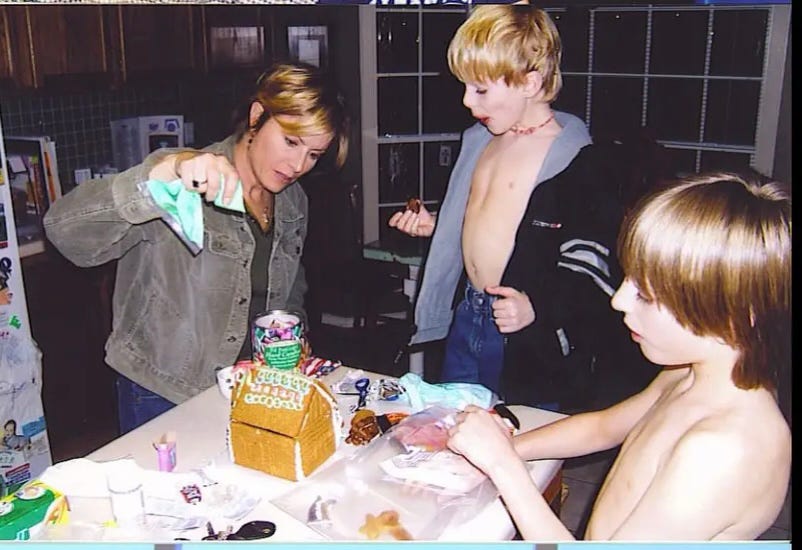 Debbie and two young boys gathering around a table with a gingerbread house on it.