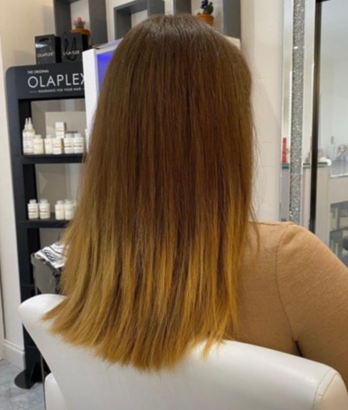 A young, white female sat in a salon. Only the back of her head is visible. She has brown, mid-back length hair. It's pin straight, and has a slightly grown-out blonde (yellow) balayage at the bottom. 