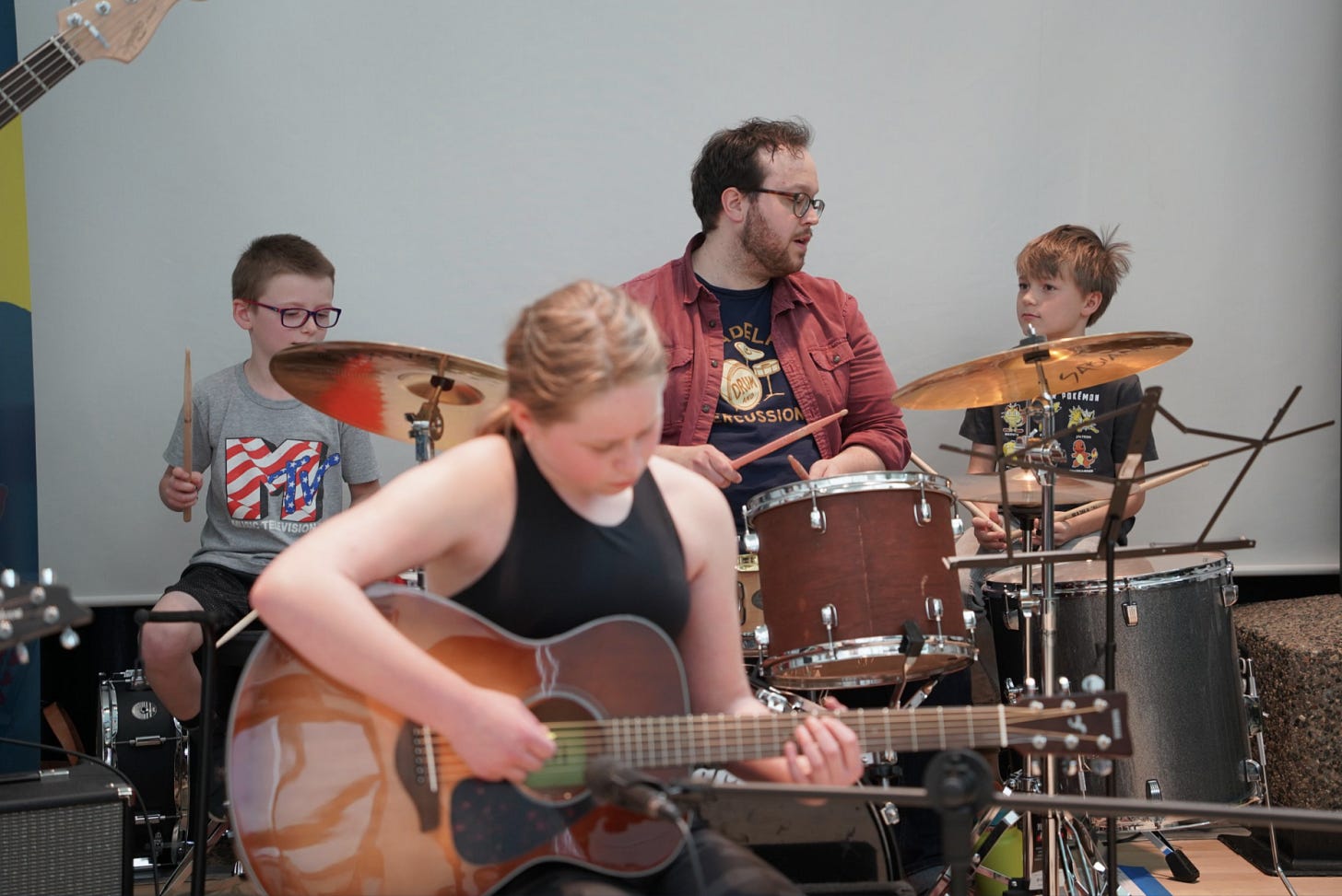 Andy performing with students at a concert. Andy is sitting on the main drum set with two younger students hitting drums on the side. Student playing acoustic guitar in front. 