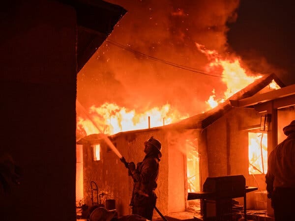 A firefighter hoses down a residential building with flames raging in the background.