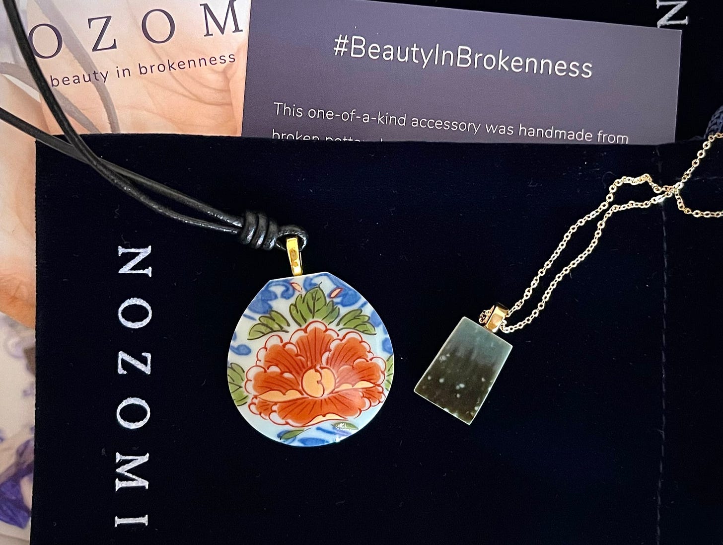 Two pendant necklaces, one featuring a floral design and the other in shades of dark gray, with Nozomi Project ephemera behind them.