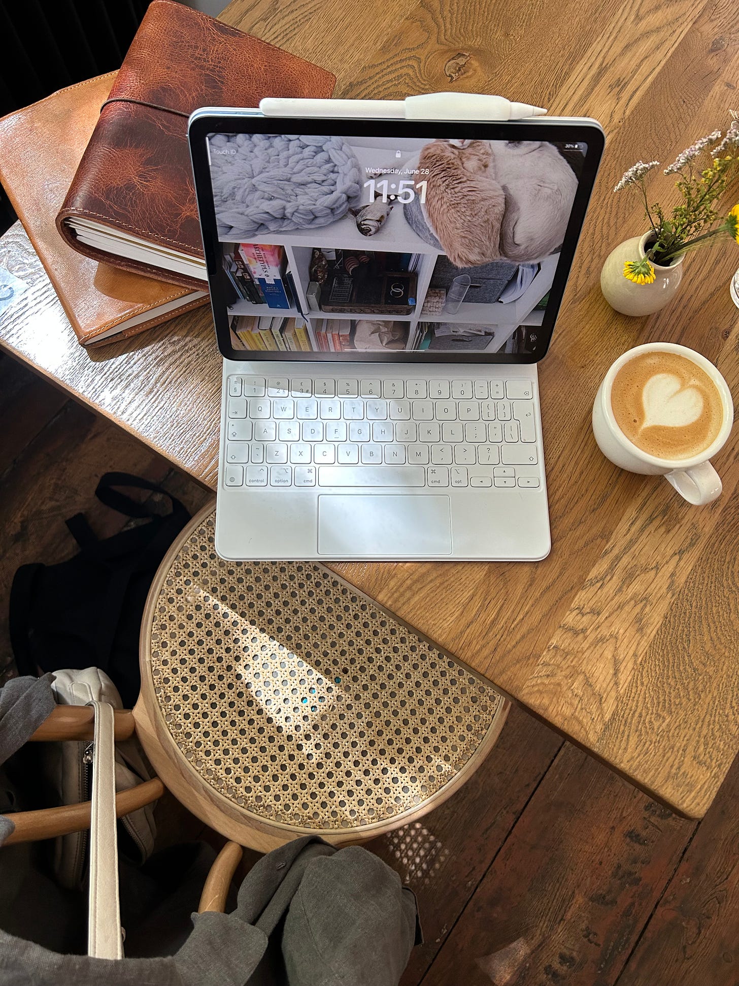 ipad with keyboard sits on a cafe table next to a cappucino, small vase of yellow and white flowers and a stack of notebooks