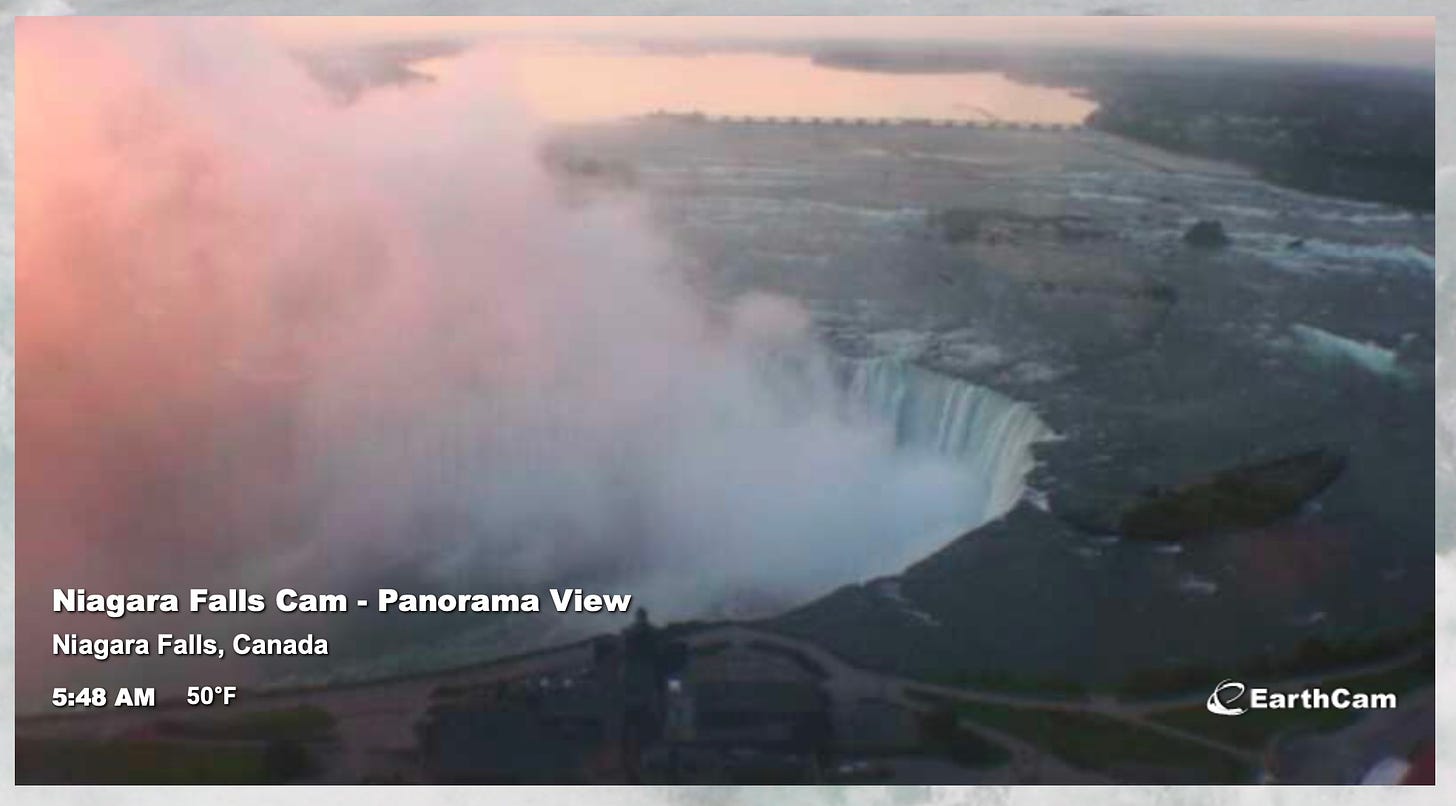 screen shot of the camera view of Niagara Falls, at sun rise with pink on the white foam of the water rising up from the top of the falls.