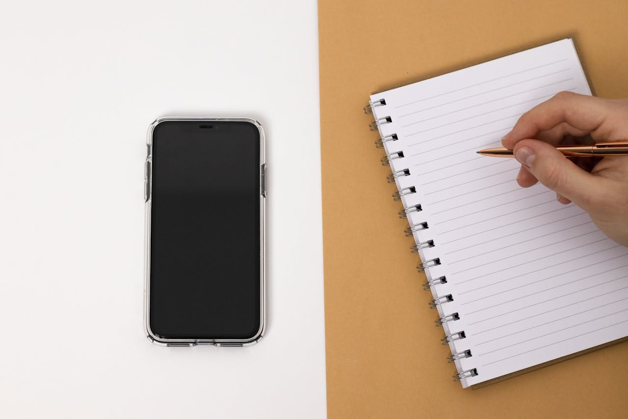 Photo by Mike Murray: https://www.pexels.com/photo/a-cell-phone-and-a-person-writing-in-a-notebook-6446248/