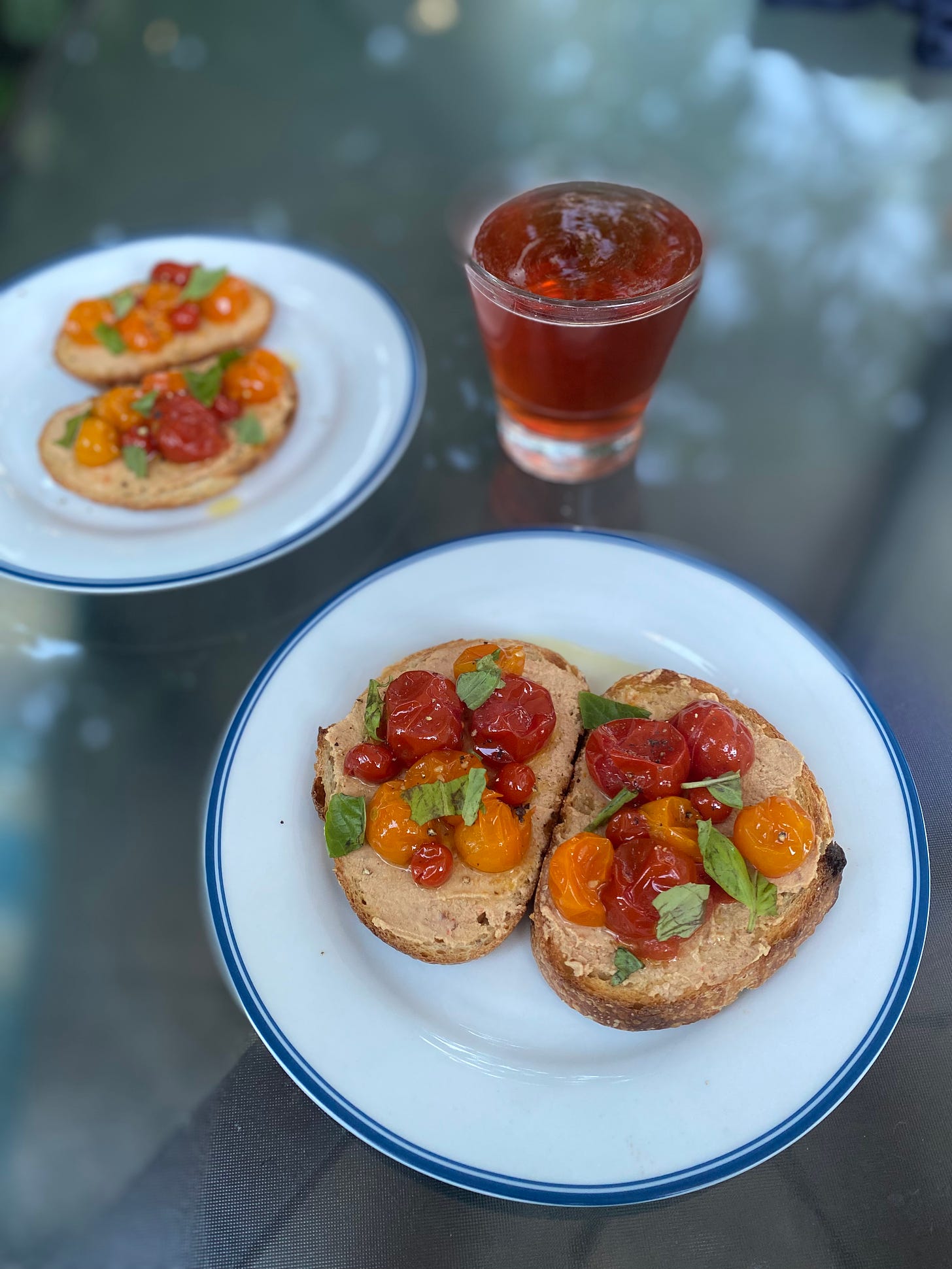 two white plates with blue rims, each with two small slices of sourdough. each piece of toast has salmon pâté, orange and red cherry tomato confit, and torn pieces of basil on top. a glass of pinkish sour beer sits between them.