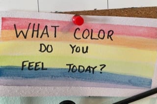 A hand made sign with a rainbow made with watercolor. Words in black ink as "What color do you feel today?"