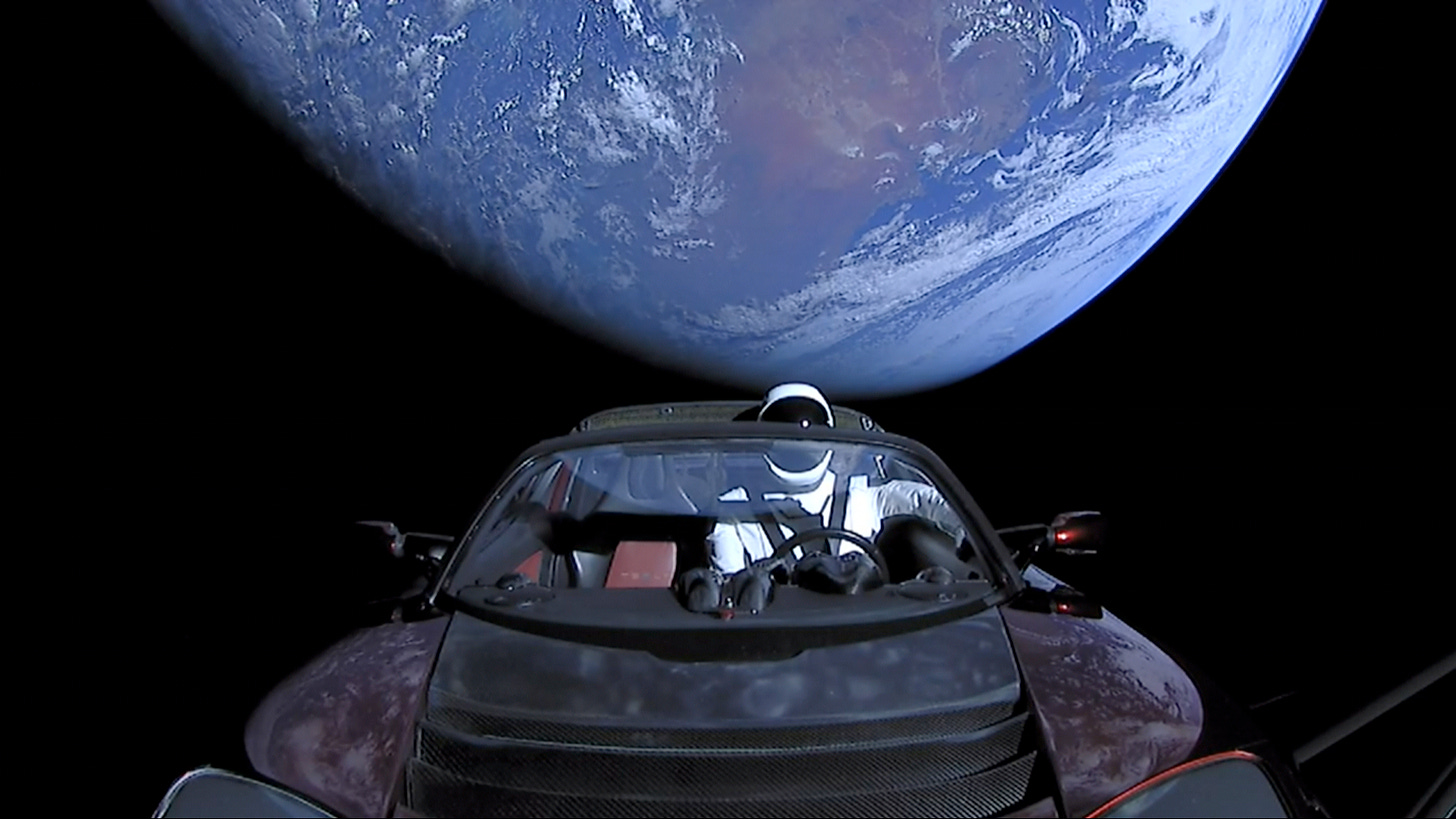 Photograph of the black emptiness of space, with planet Earth partly in shadow in the background. In the foreground is an open-top red convertible sports car, viewed from the front over the hood, with a mannequin in the driving seat that is wearing a white-and-black spacesuit