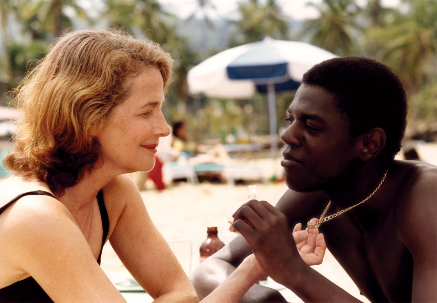 An older white woman leans closer to a younger Black man on a beach as she fondles a gold necklace around his neck