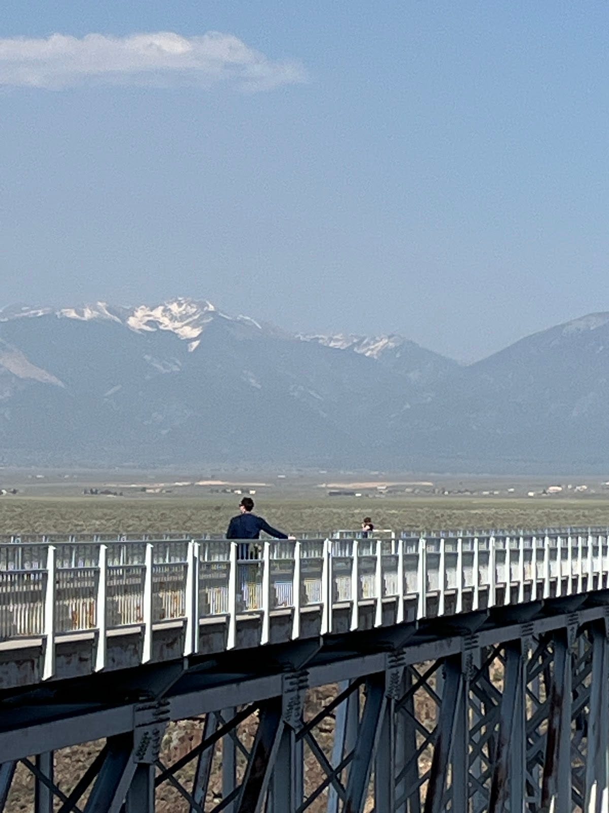 a bridge over the Rio Grande River Gorge, with two people standing on it. Mountains rise in the distance.